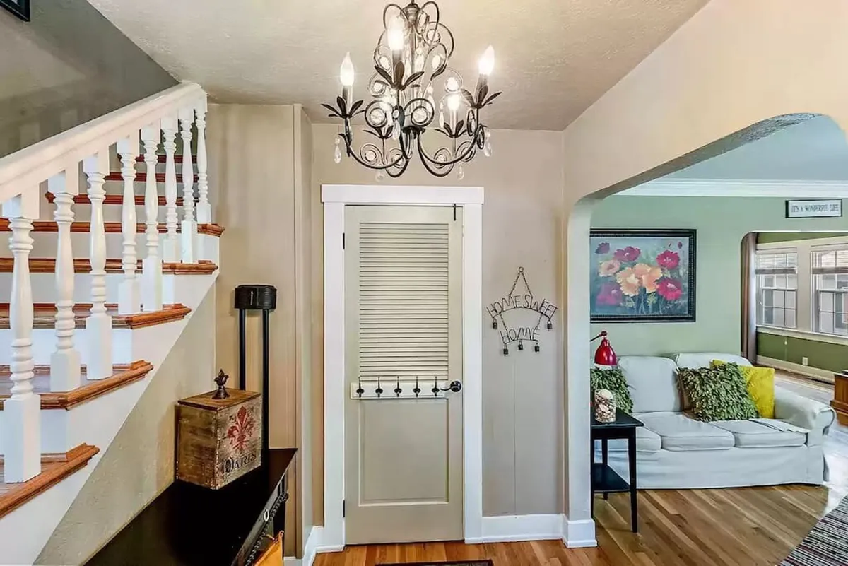 This image centers on an elegant living area featuring a decorative staircase, a sparkling chandelier, a stylish door, and a cozy couch with colorful pillows. The arrangement suggests a welcoming entry point and an open space that could easily serve as a gathering area before or after exploring the neighborhood. The period charm of the staircase and chandelier highlights the historic character of the home.