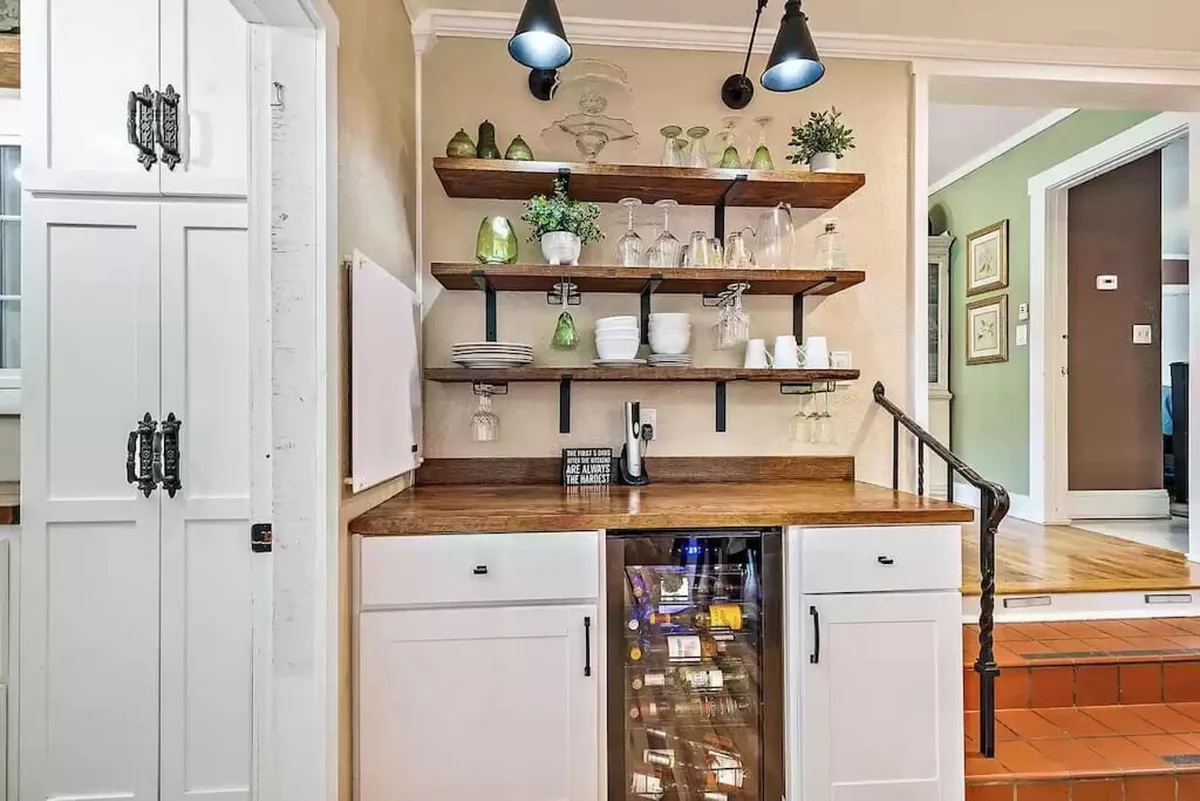 A dedicated wine bar area featuring a wine cooler, glassware neatly displayed on shelves, decorative plants, and a warm wooden countertop. The ambiance suggests a space ideal for entertaining and sharing drinks before or after a meal.