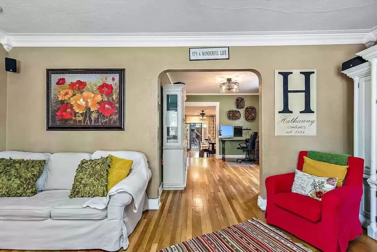 A sunny, cheerful living room with a plush sofa adorned with colorful cushions, a vibrant red armchair, floral artwork, and an open archway. The space feels lively and sociable, perfect for relaxing between cultural adventures or planning the next walk through historic Medford.