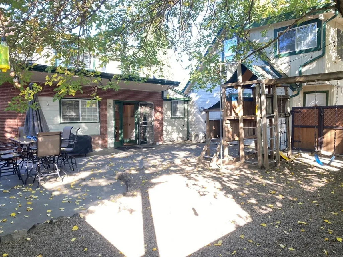 This image presents a vibrant backyard scene featuring a dedicated playground structure, an outdoor dining set, and lush greenery. The back door entrance is also visible, seamlessly connecting indoor and outdoor living. The mood is lively and inviting, with space for both active play and family meals in a welcoming natural environment.