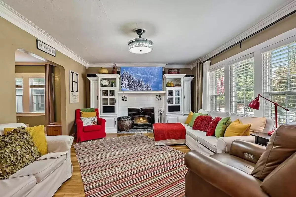 A warmly lit living room featuring a classic wood-burning fireplace, a smart TV above the mantel, a plush sofa with colorful cushions, and a cozy rug. The seating is arranged to promote conversation and relaxation, perfectly embodying the inviting atmosphere of the main gathering space.
