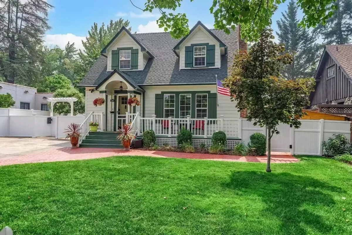 A welcoming front porch overlooks a green, well-kept lawn dotted with vibrant flower pots and an American flag. The scene hints at a relaxing outdoor space perfect for gatherings or unwinding while children play nearby.