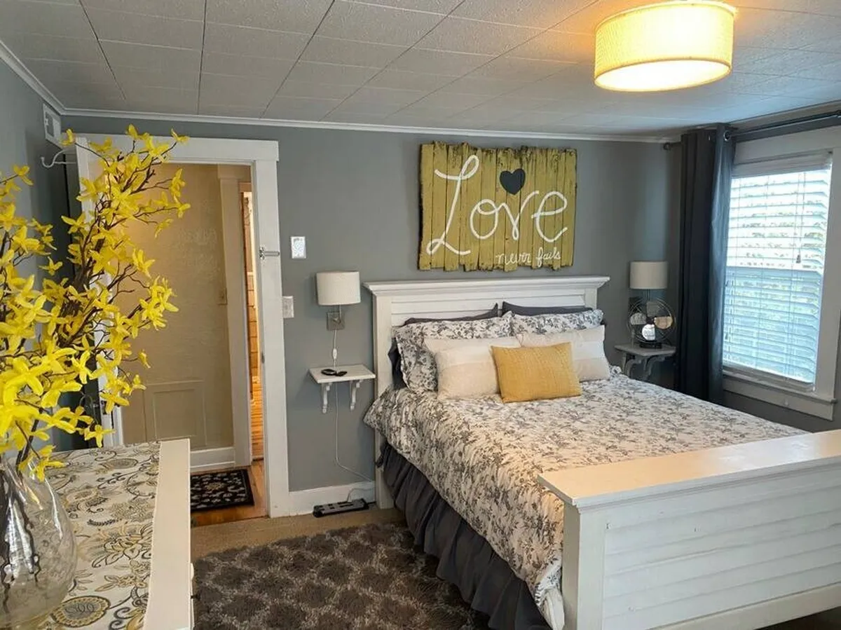 A tranquil bedroom with a comfortable bed featuring decorative pillows, a 'Love' sign on the wall, and a touch of freshness with a yellow flower arrangement. Sunlight streams through the window, illuminating a cozy space perfect for relaxation.