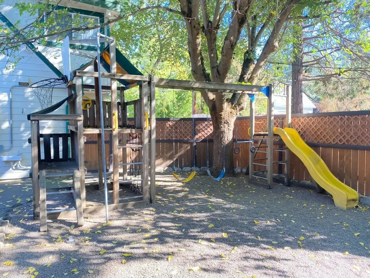The image showcases a classic playground structure with a slide and swing set tucked under a mature tree. The open space invites children to play and families to gather for outdoor picnics or celebrations. The setting is filled with natural light, emphasizing a joyful and active atmosphere.