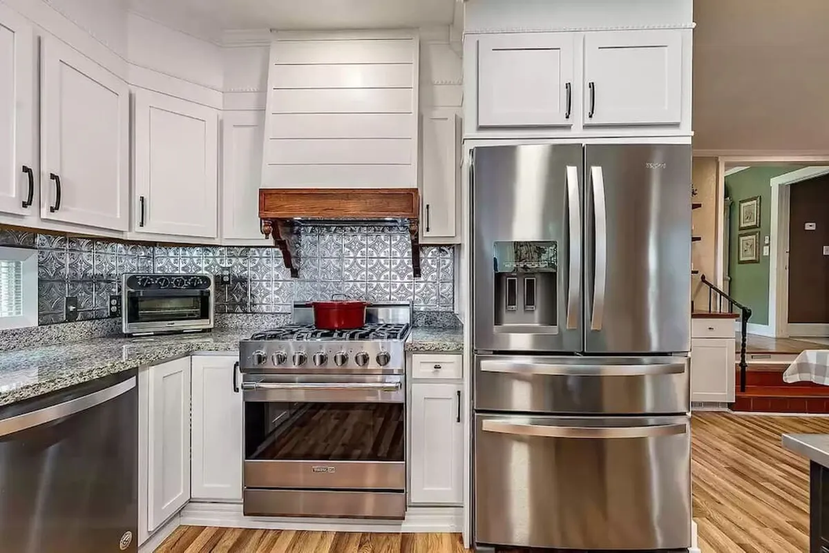 A modern kitchen with a stainless steel refrigerator, granite countertop, sleek stove, and decorative backsplash. A red cooking pot sits on the stovetop, suggesting recent meal preparation. The space feels open and inviting, with enough room for family cooking sessions or entertaining guests.
